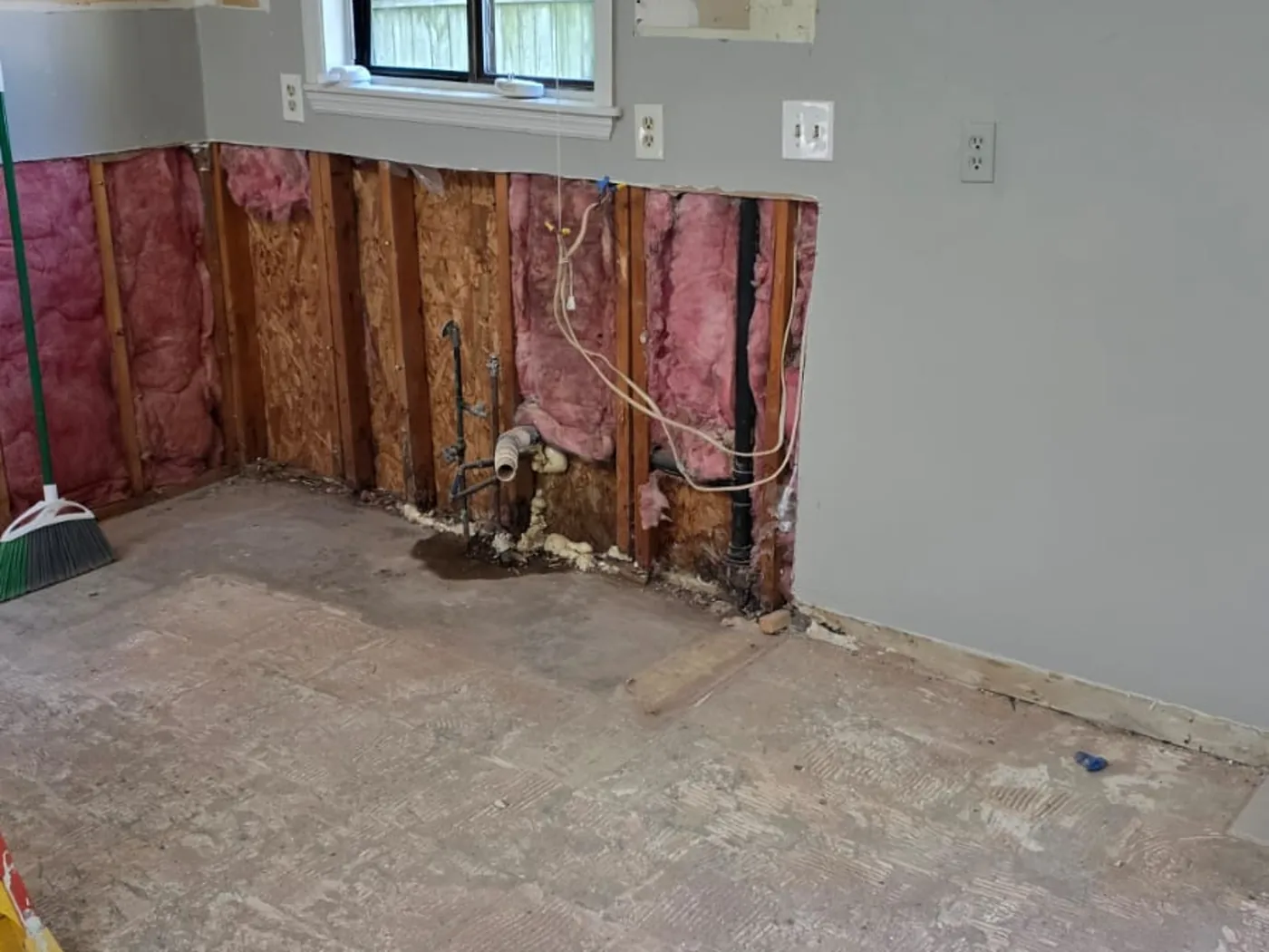 Kitchen stripped back to the studs during demolition, with insulation and plumbing exposed along the sink wall and the old tile floor removed.