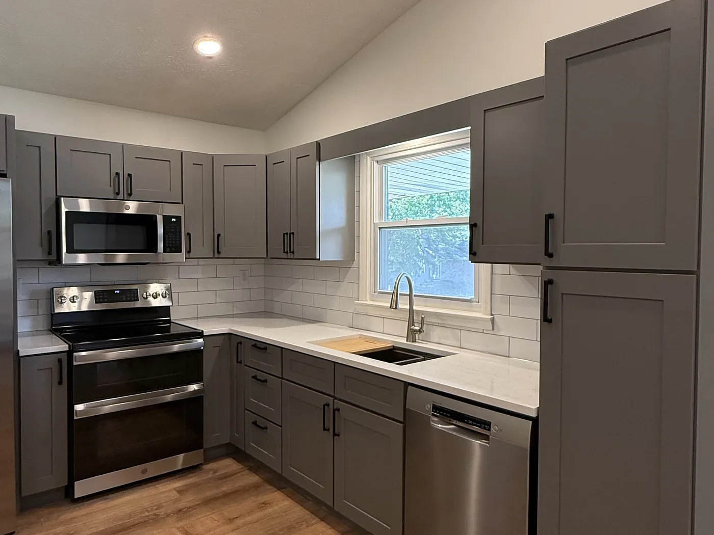 The finished kitchen with gray shaker cabinetry, quartz counters, a white subway tile backsplash, stainless range, microwave and dishwasher, and new wood-look flooring.