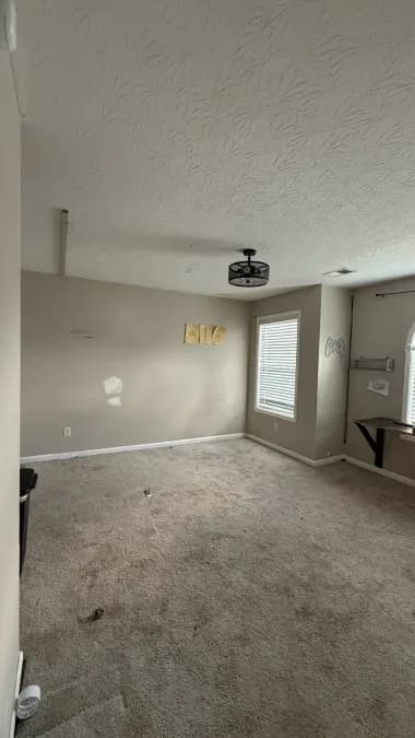 Carpeted room with wall patches, a ceiling light, and windows before flooring replacement.